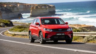 Red Geely EX5 compact SUV driving on the Great Ocean Road along the Australian coast with the Twelve Apostles in the background
