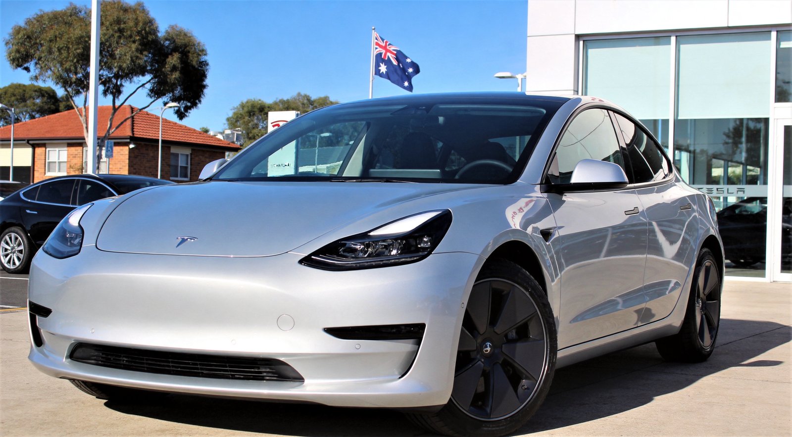 Silver Tesla Model 3 sedan parked at an Australian Tesla dealership showroom with an Australian flag flying on the pole behind
